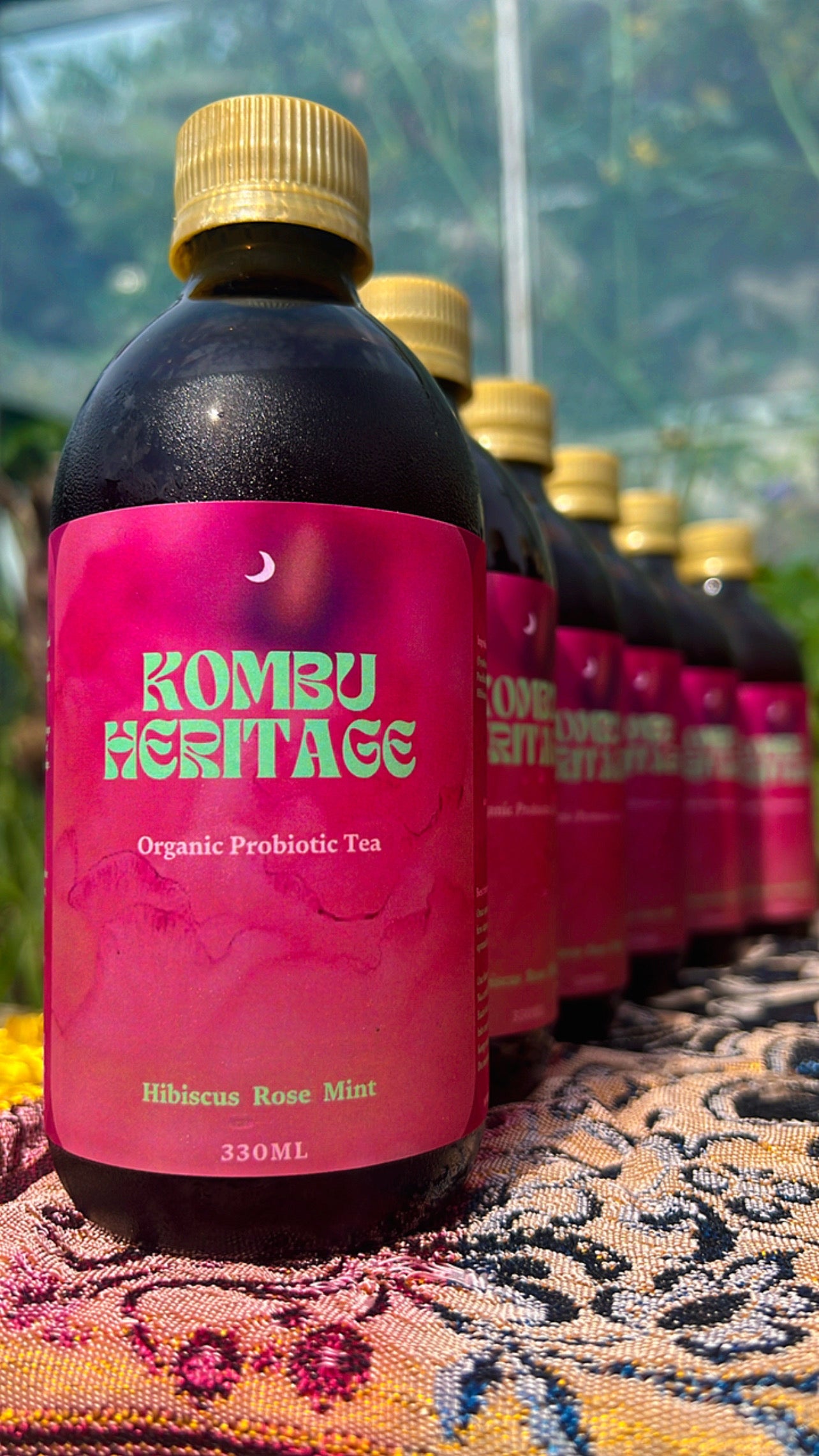 A row of bottles labeled "Effervescent Explorer 24X" from KombuHeritage stands on a patterned cloth. The label indicates it's an organic, probiotic kombucha tea with a hibiscus rose mint flavor. The dark bottles with golden caps and pink labels sit against a backdrop of greenery, suggesting an outdoor setting perfect for an Effervescent Explorer.